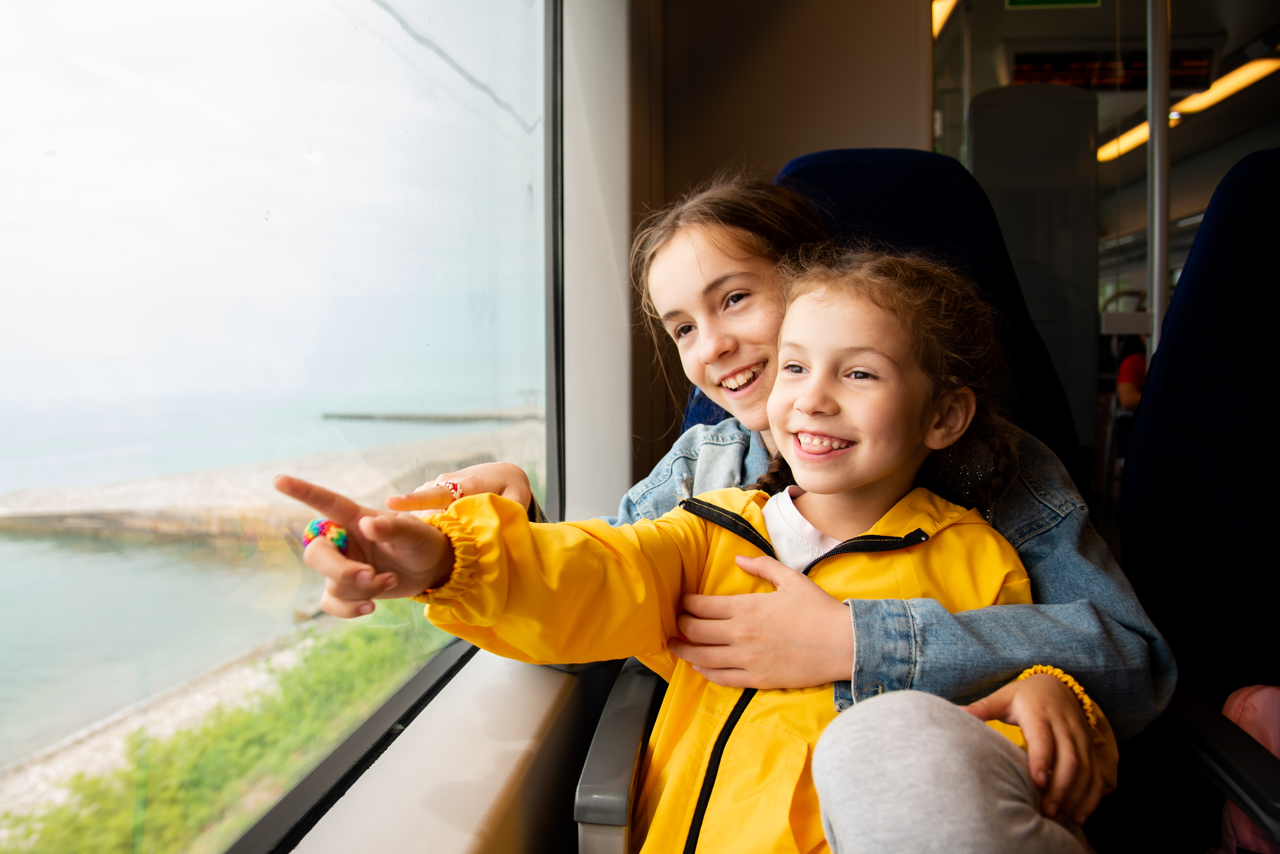 Zwei weiblich gelesene Kinder sitzen gemeinsam auf einem Sitzplatz im Zug. Das jüngere Kind sitzt auf dem Schoß des älteren.
