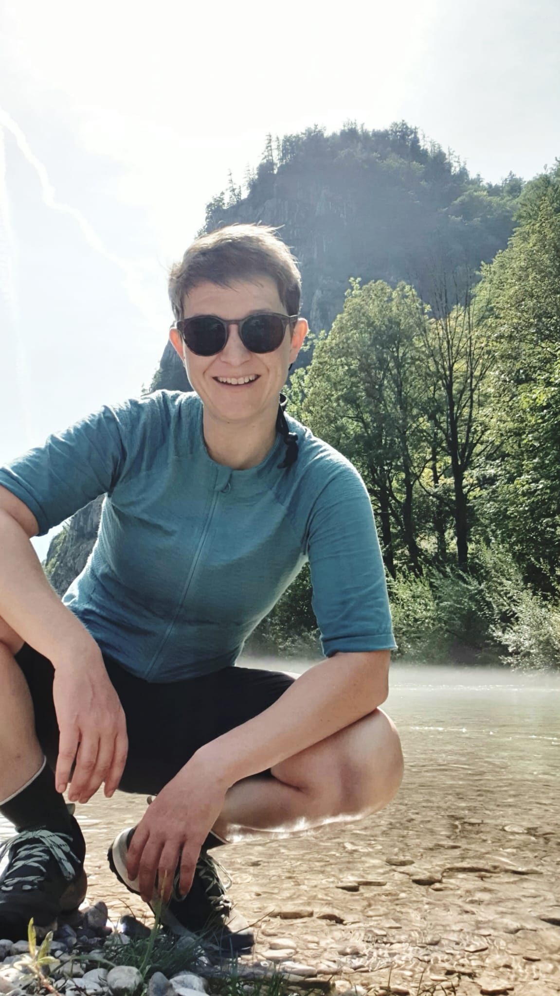 Katrin vom Team cyclbee im Fahrrad-Outfit in der Hocke an einem Bach-Ufer. Im Hintergrund ein Berg.